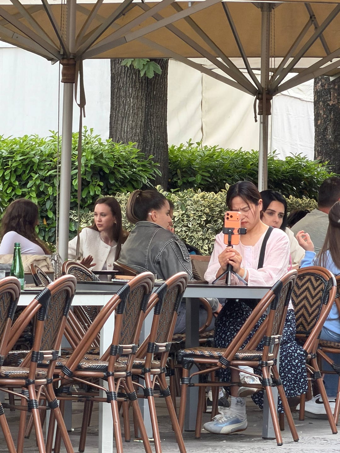 A person at a Belgrade café terrace holding a phone on a small tripod, one lens facing out toward the tables.