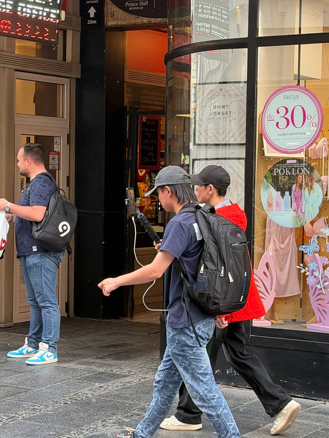 A person walking through a Belgrade shopping street holding a phone on a gimbal, backpack on.