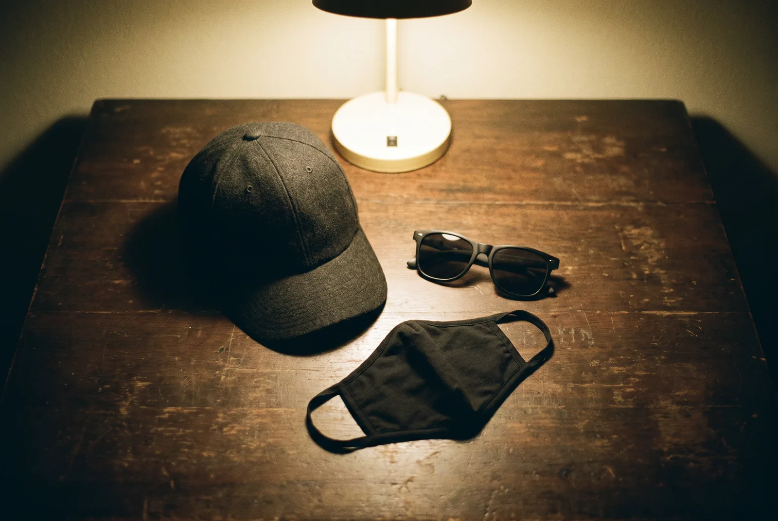 A baseball cap, dark sunglasses and a folded black face mask laid out on a wooden table under a desk lamp, photographed from above.
