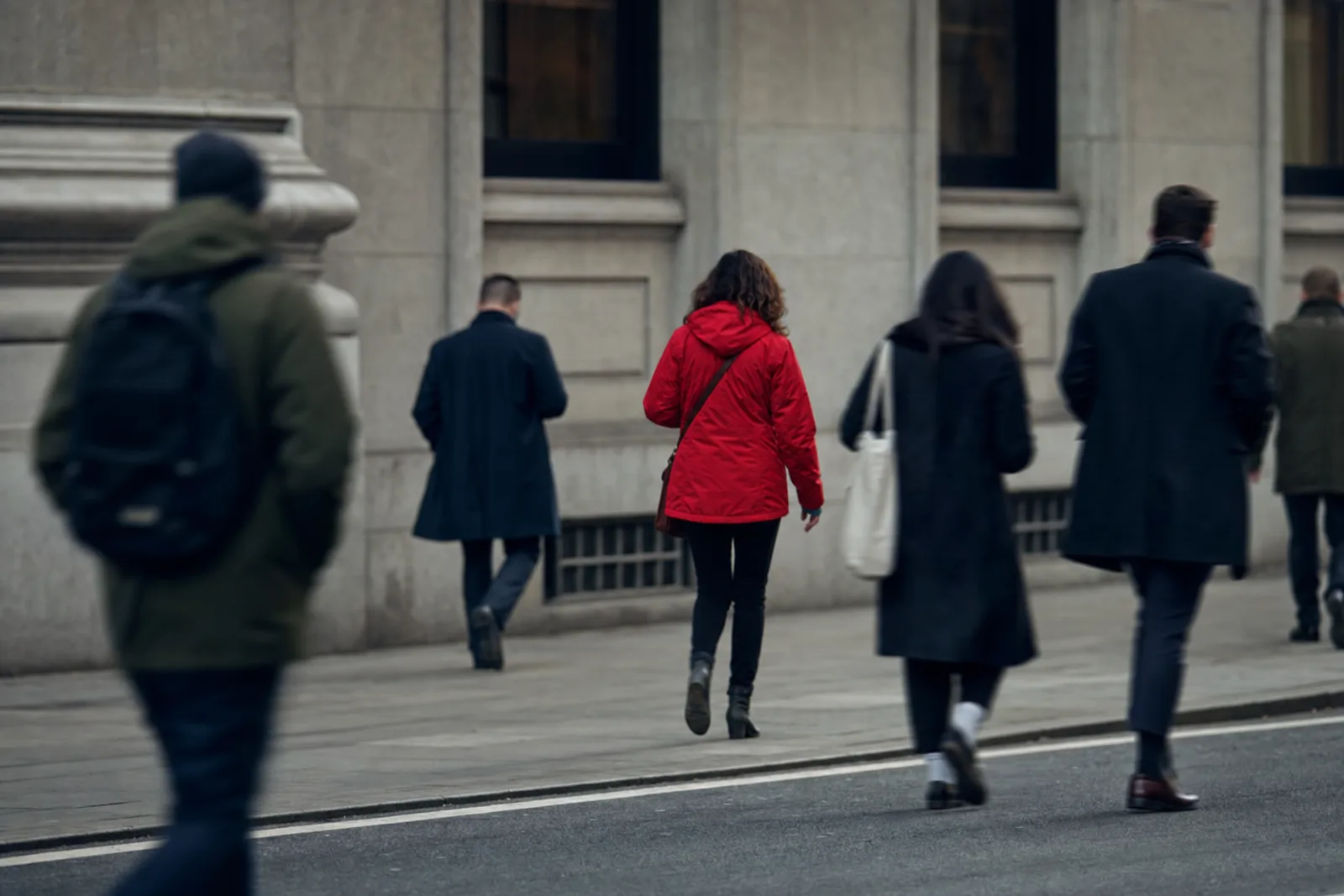 A pavement seen from across the road: several anonymous pedestrians walking away from the camera, all motion-blurred and indistinct, except for one person in a bright red jacket who is rendered sharply and stands out from everyone around them.