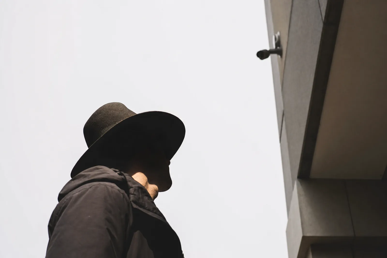 Looking up from pavement level at a person in a wide-brimmed hat and dark hooded jacket against a bright blank sky, with a small dark camera housing mounted high on the corner of a building nearby. The brim throws a hard shadow that fills the face with black, leaving only the jaw and neck lit.