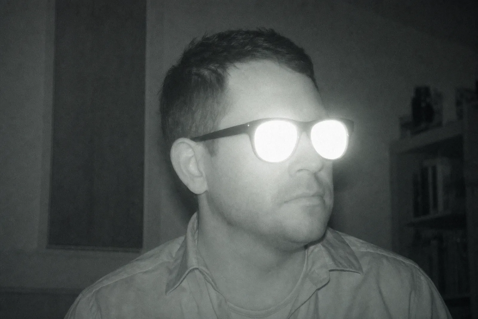 An infrared security-camera view of a man in ordinary dark glasses. Both lenses blaze back as hard white discs of reflected IR, wiping out the entire eye region, while the rest of his face and shirt are normally exposed.