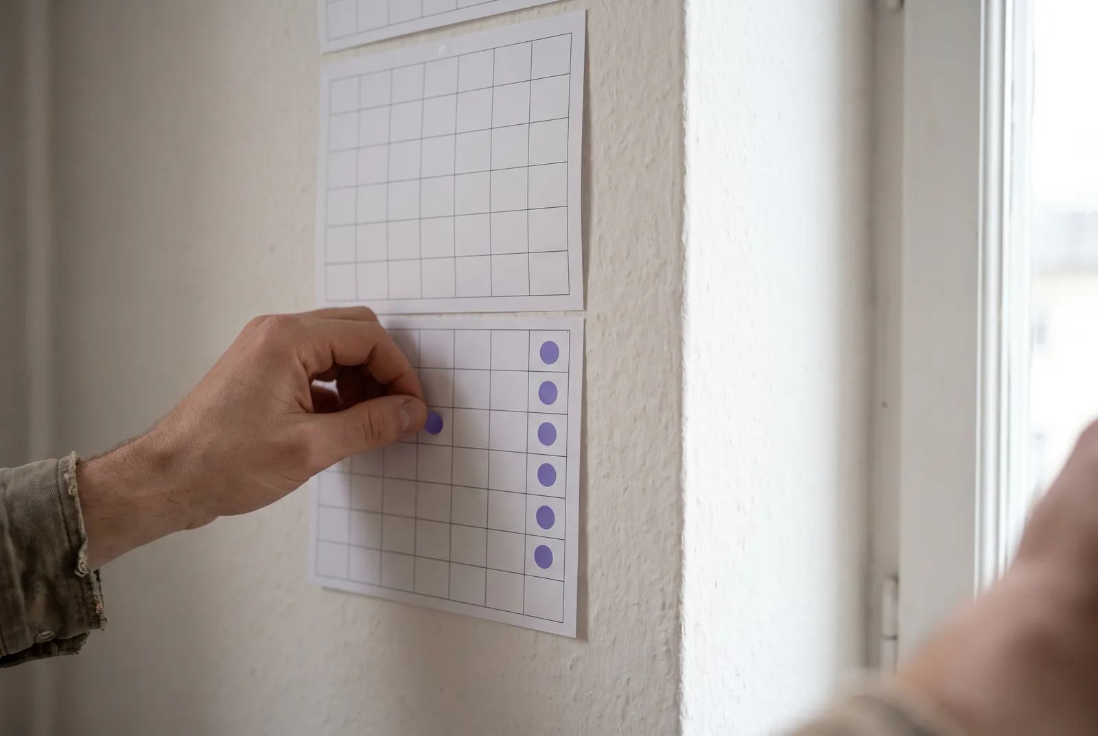 A wall calendar of blank grid squares with a small violet sticker placed on one square each month, forming a repeating line. A hand presses on the newest one.